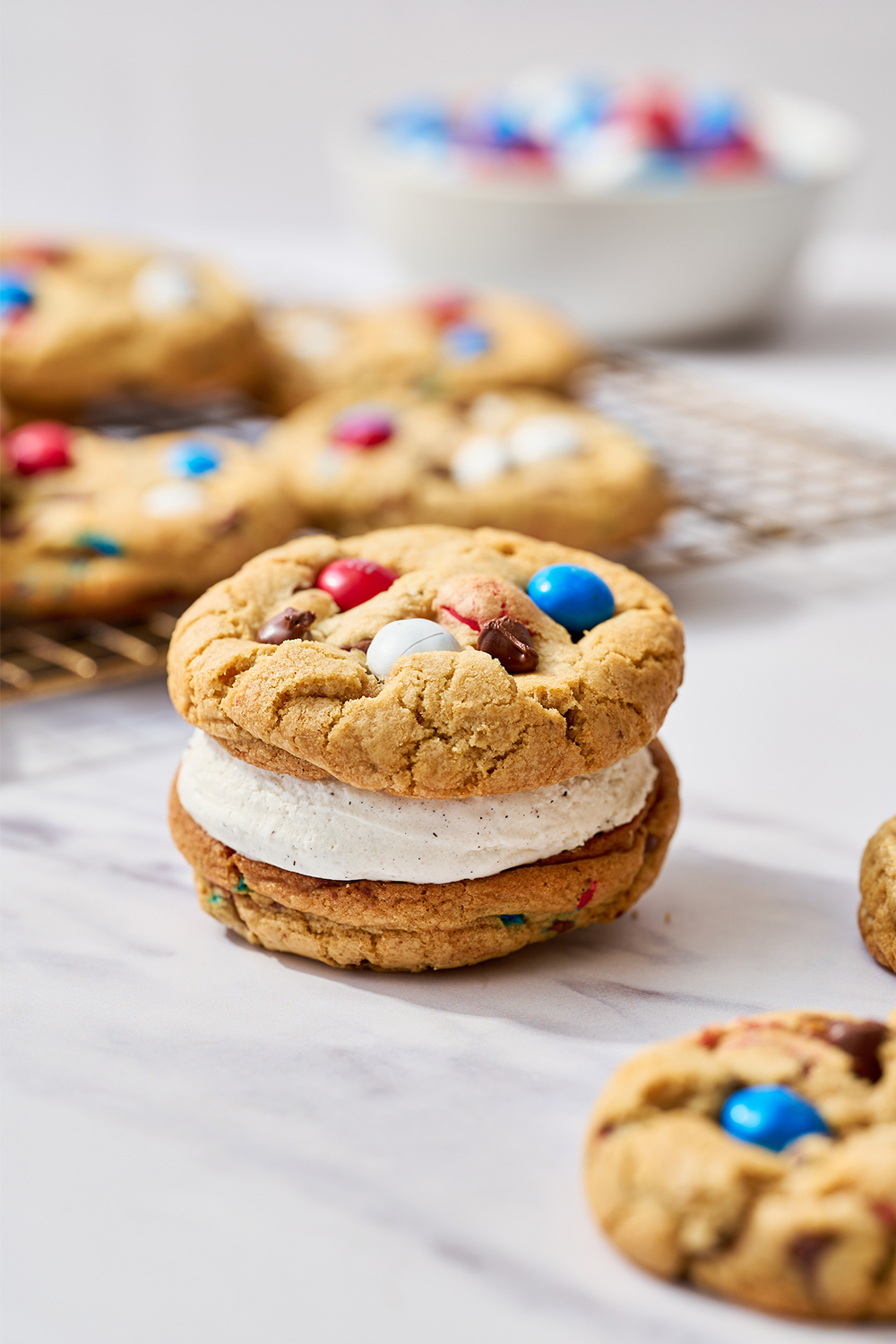 4th of July ice cream sandwich, sitting in front of a wire rack full of red, white, and blue M&M-studded chocolate chip cookies
