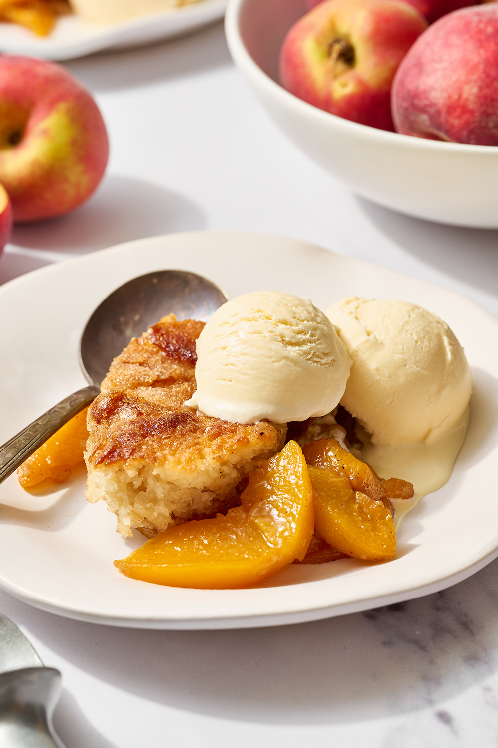 warm peach cobbler served with vanilla ice cream, on a white ceramic plate with fresh peaches in the packground