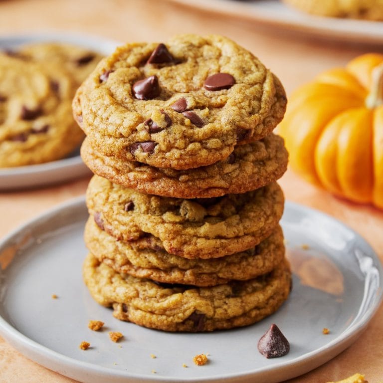 several Brown Butter Pumpkin Chocolate Chip Cookies stacked on a white plate, with a small pumpkin behind.