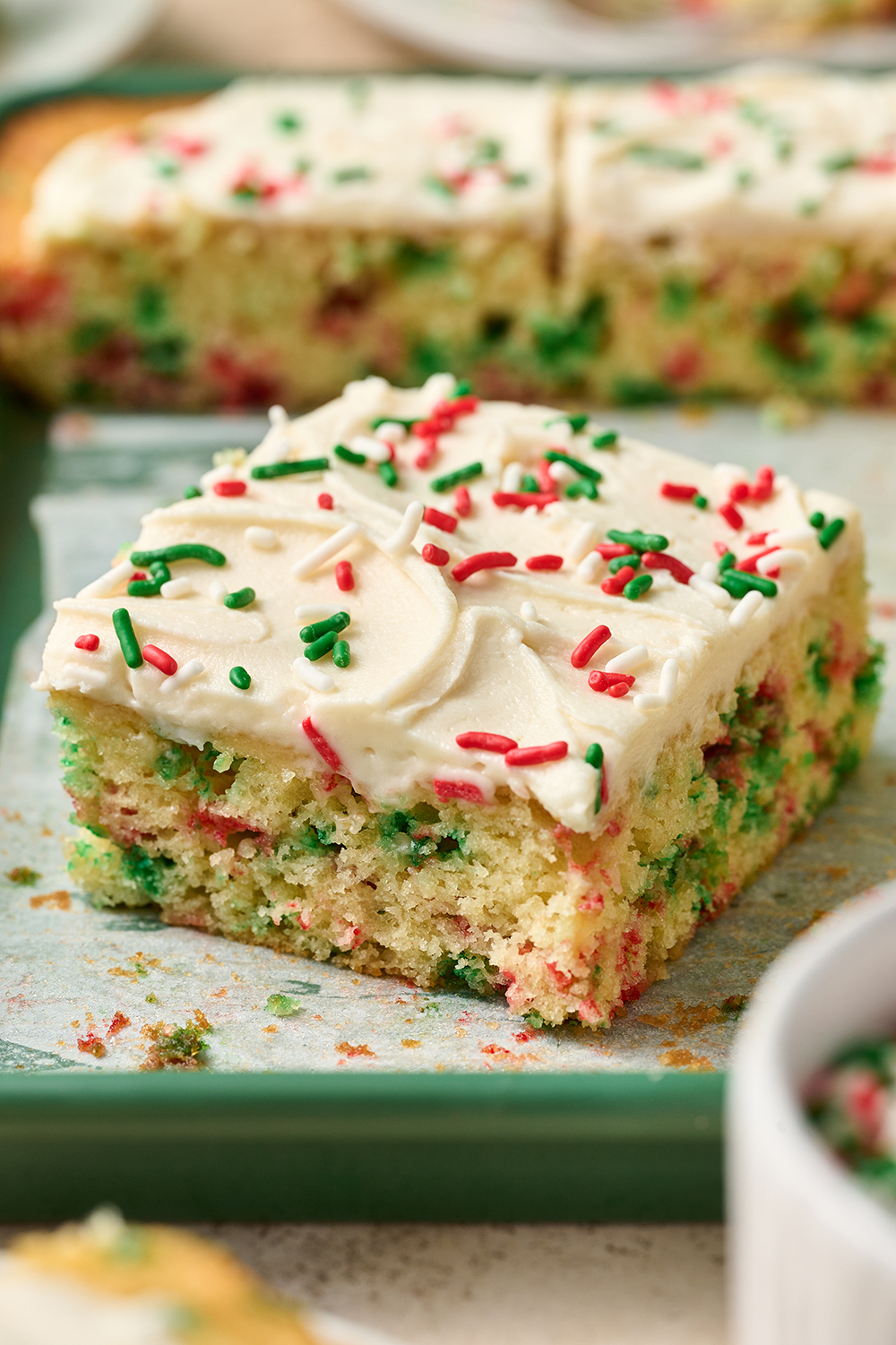 the cake in the background is all sliced up, with one slice close to the front, frosted and topped with holiday sprinkles.