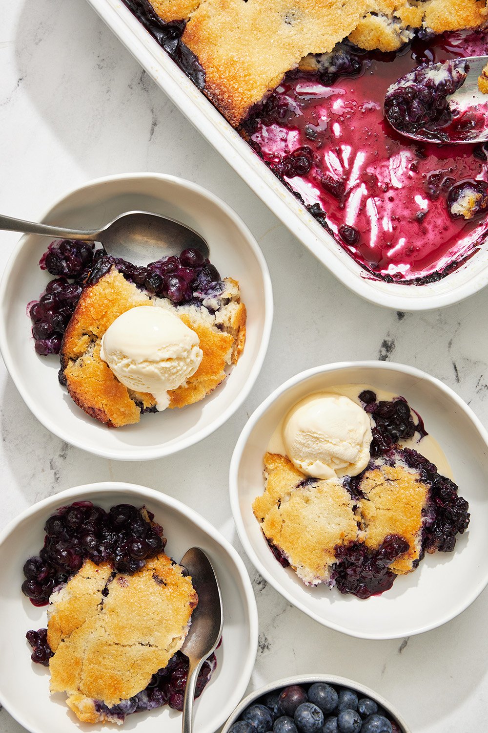 bowls of blueberry cobbler with vanilla ice cream next to a baking dish