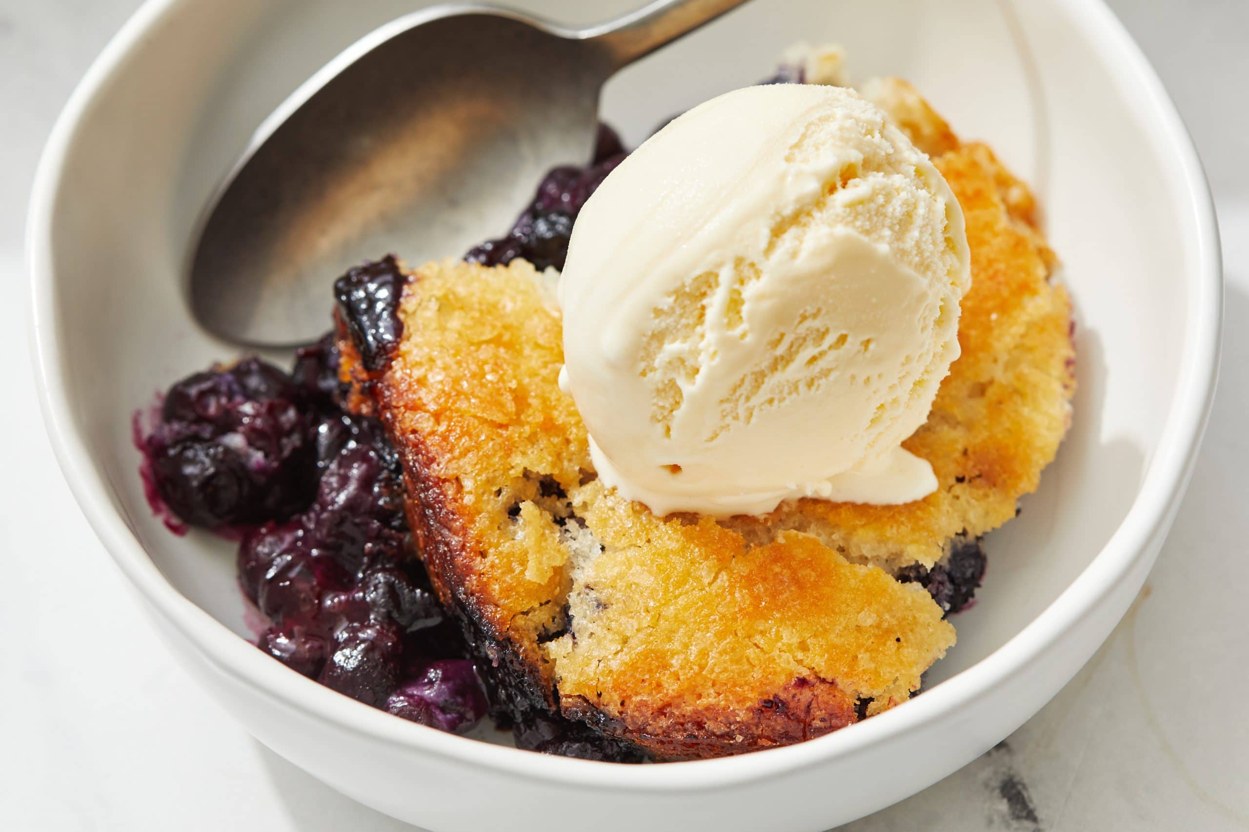 Close up of white bowl holding warm bluberry cobbler, a scoop of vanilla ice cream, and a spoon