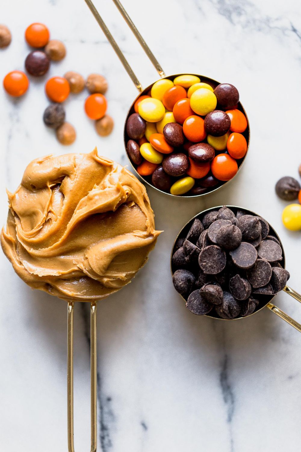 three measuring cups, one with peanut butter, one with Reese's Pieces, and one with chocolate chips, ready to be made into cookies.