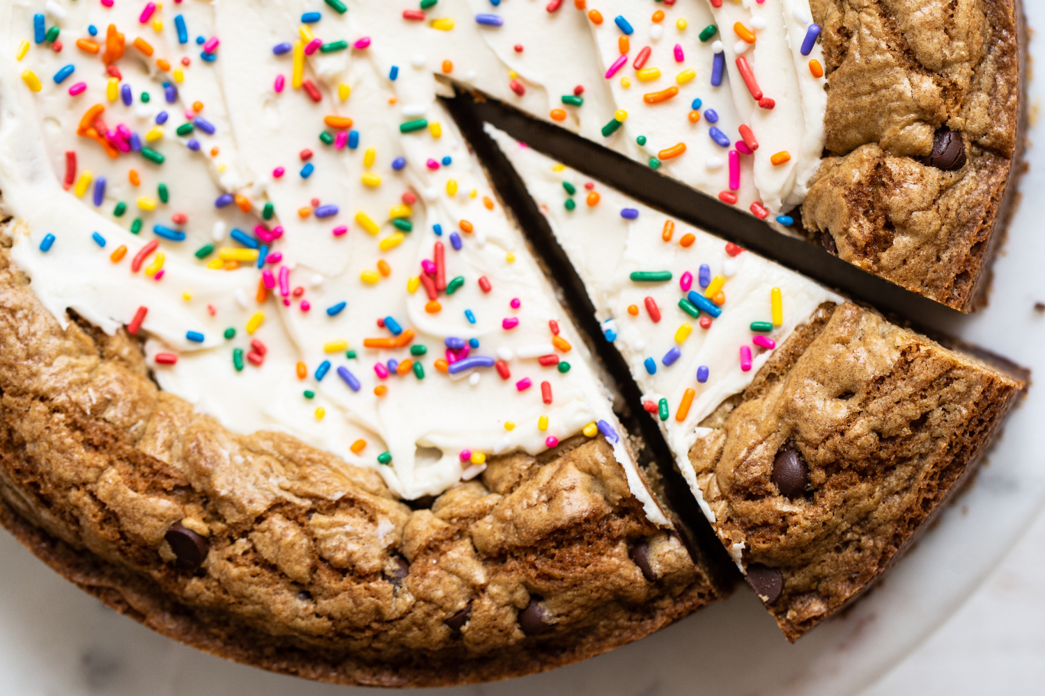 chocolate chip cookie cake with creamy white frosting and rainbow sprinkles, with a slice being cut to serve.