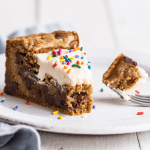 a slice of thick, gooey cookie cake with white frosting on a plate.