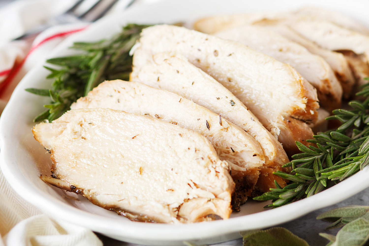 Sliced slow cooker turkey breast, on a plate, on top of a bed of fresh rosemary, with a fork ready to serve guests.