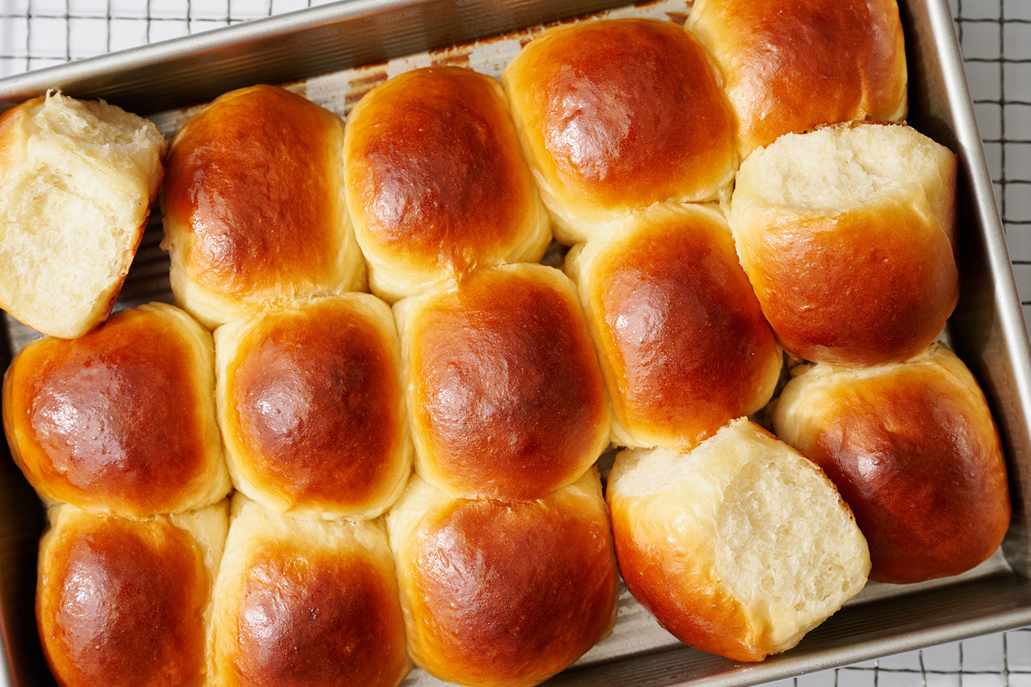 homemade hawaiian bread rolls with butter brushed on top, some still in the pan and a couple removed and turned on their side to show their fluffy texture.