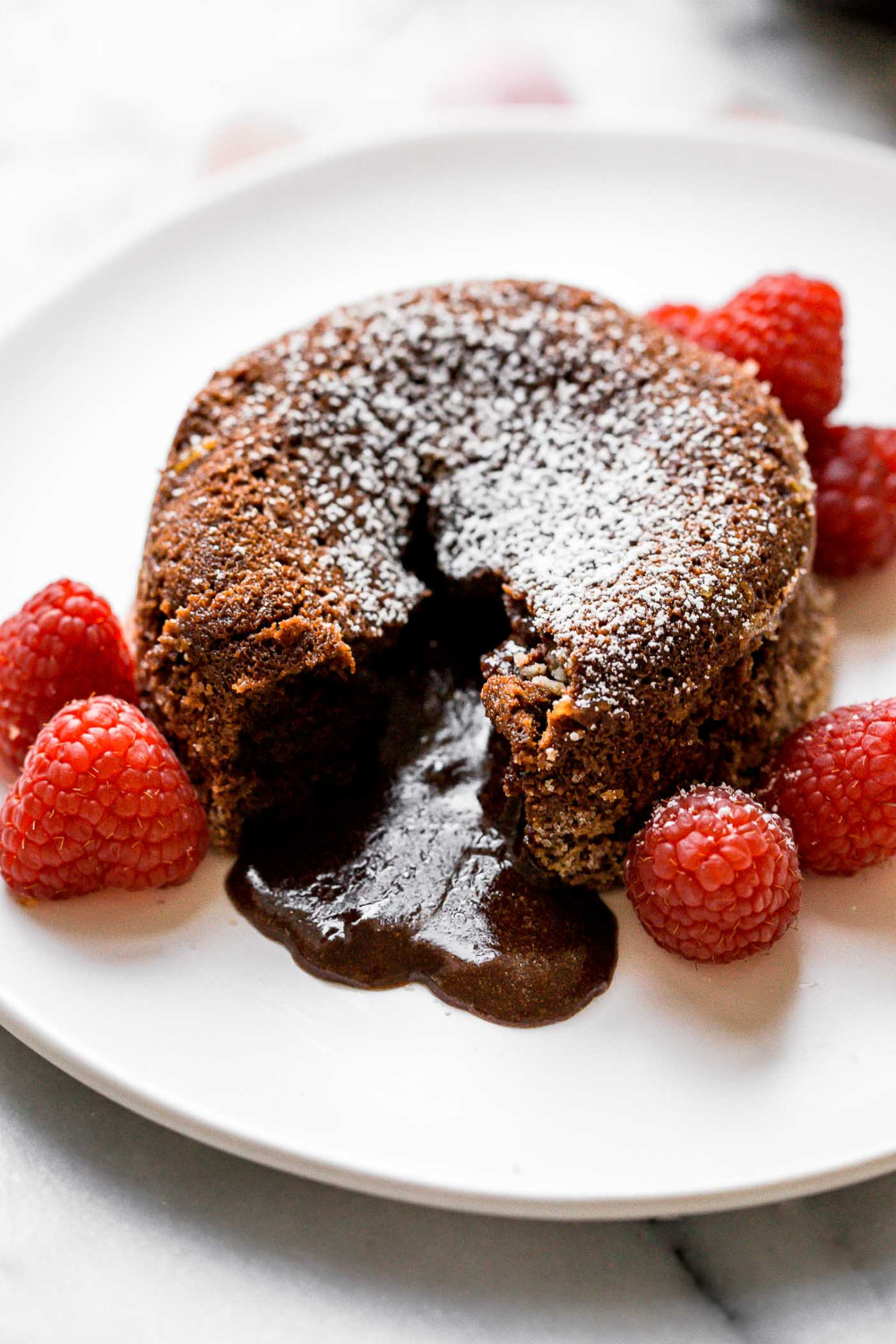 a chocolate lava cake on a white plate, surrounded by fresh raspberries, with a bite taken out so you can see the molten interior flowing out.