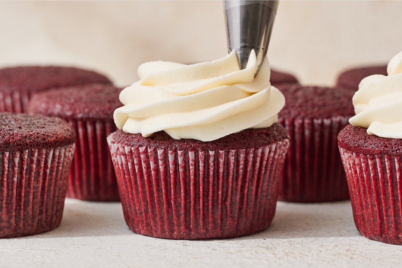 piping cream cheese frosting onto a red velvet cupcake
