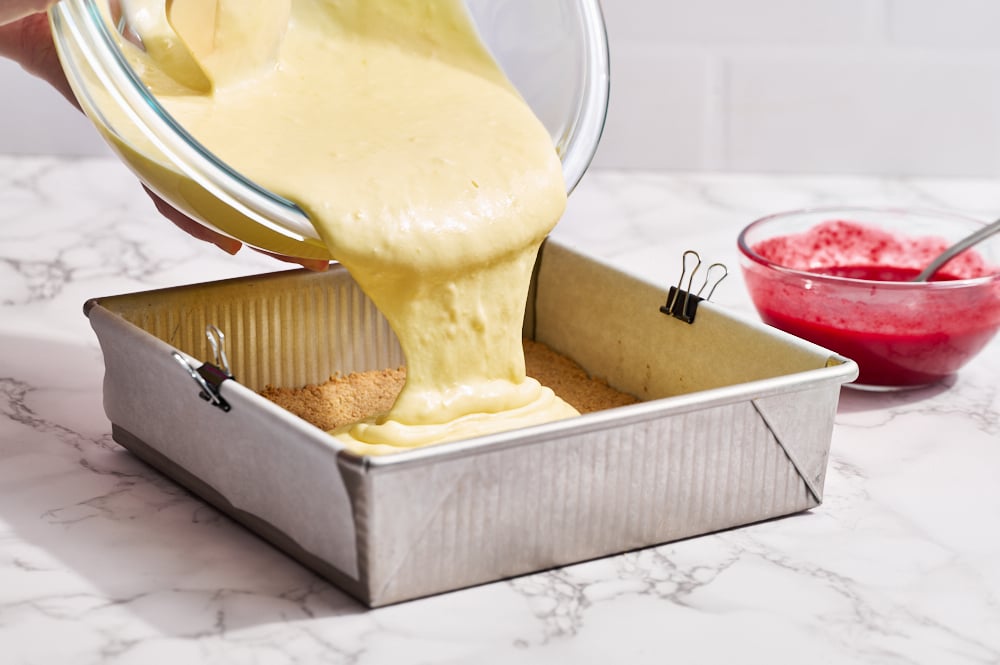 cheesecake batter being poured into a square metal baking pan