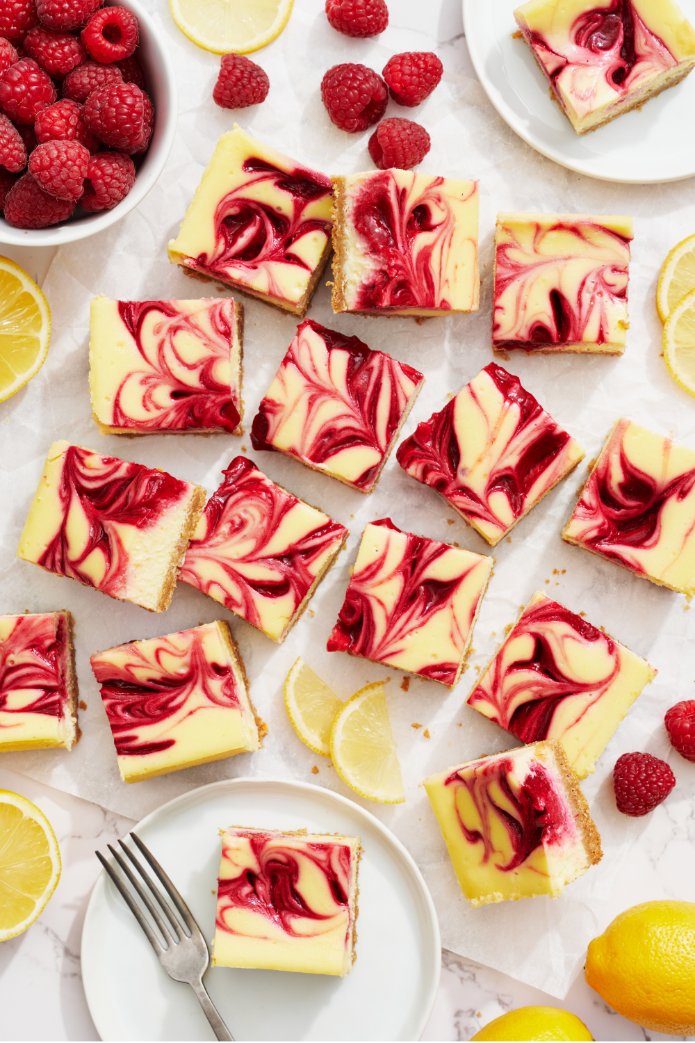 the whole pan of raspberry cheesecake bars on a white countertop, with fresh lemons nearby and a bowl of fresh raspberries.