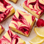 slices of raspberry lemonade cheesecake bars with a few fresh raspberries and slices of lemon nearby.