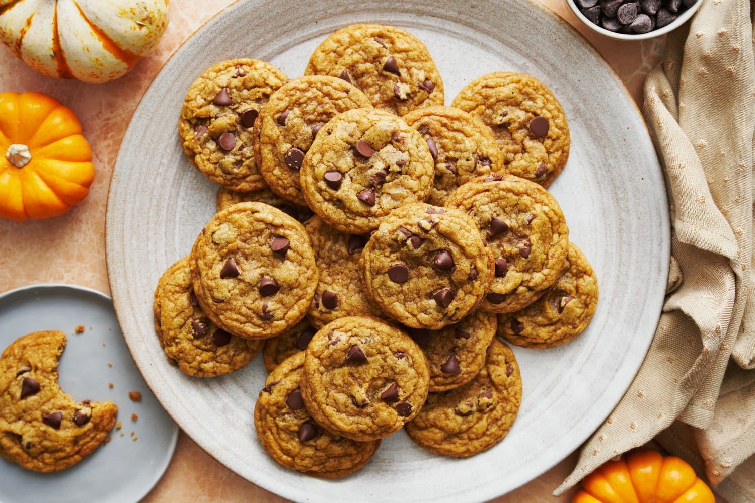 overhead of white platter with brown butter pumpkin chocolate chip cookies piled on top, with a half bitten cookie on a plate nearby