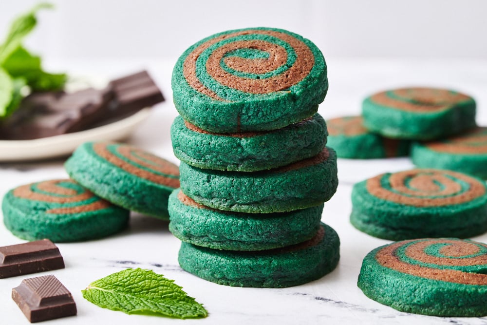 a stack of pinwheel cookies, with more cookies and some chocolate and fresh mint in the background.