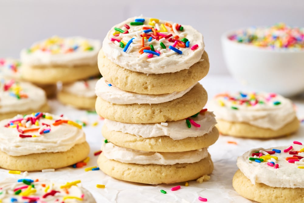 a stack of iced cookies with sprinkles, with more cookies and a bowl of sprinkles in the background.