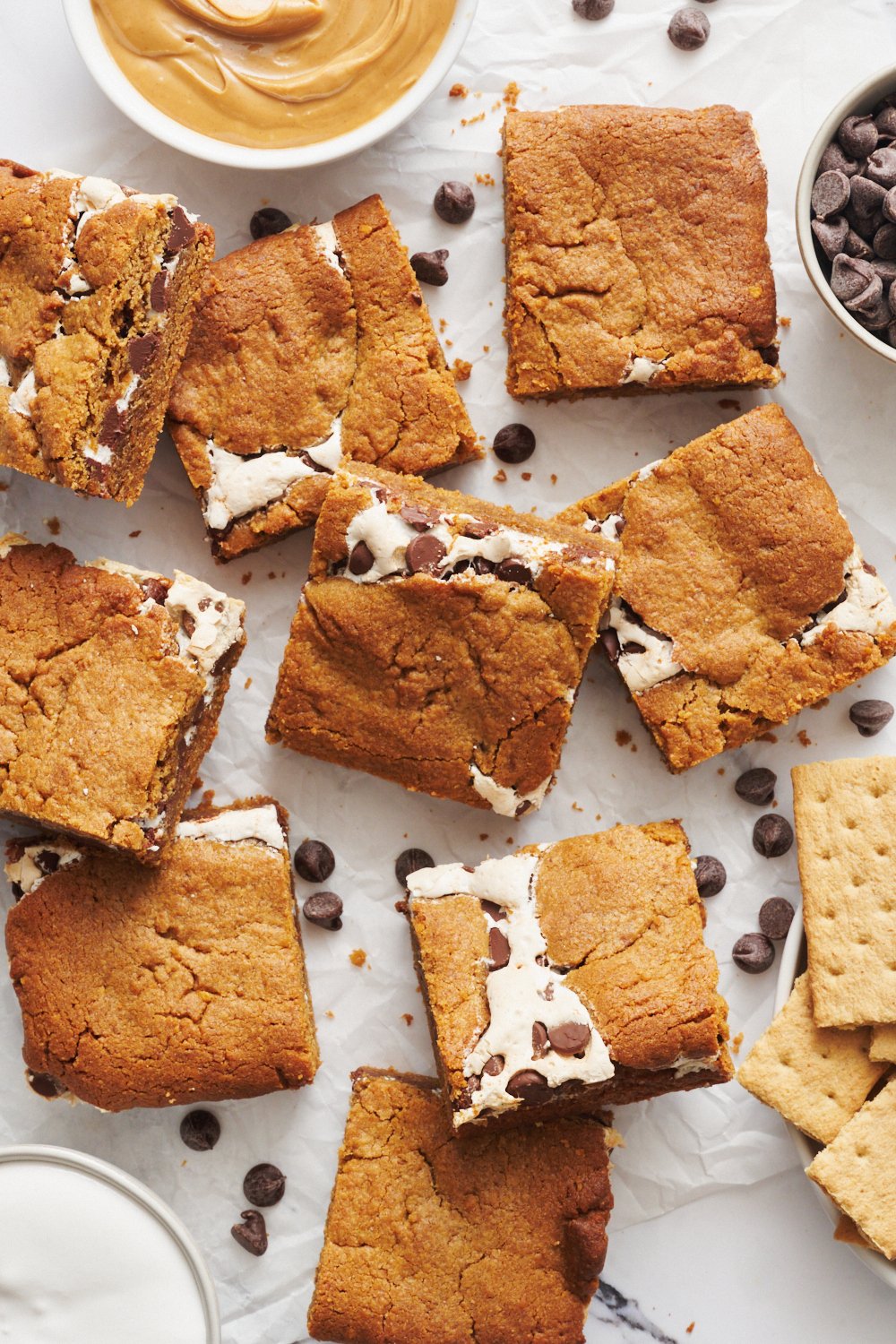 several slices of PB s'mores blondies with a bowl of peanut butter and a bowl of marshmallow creme nearby, with chocolate chips scattered throughout.