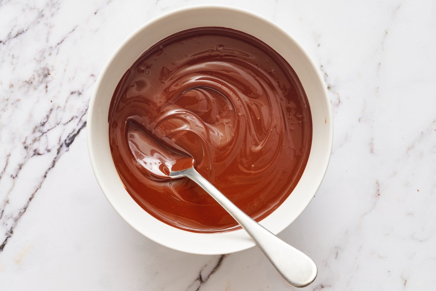 a small bowl of melted chocolate with a spoon on a marble surface.