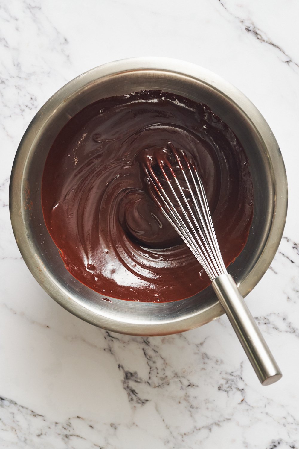 a bowl of Chocolate Ganache with a whisk in it, on a marble surface.