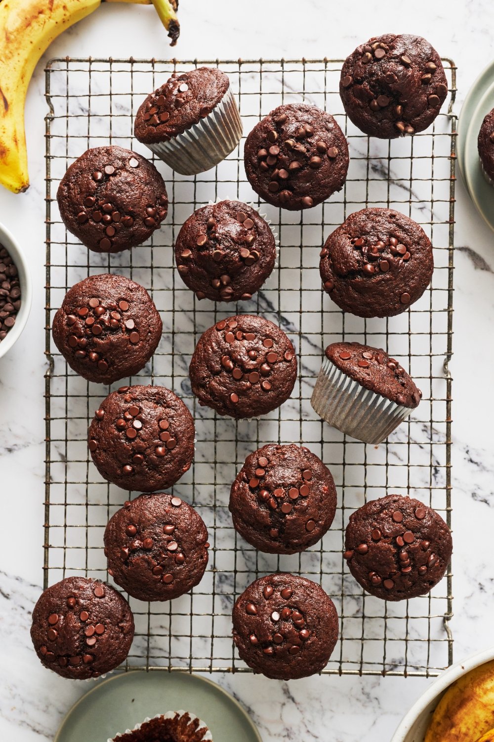 a batch of double chocolate banana muffins on a wire rack with some bananas nearby.