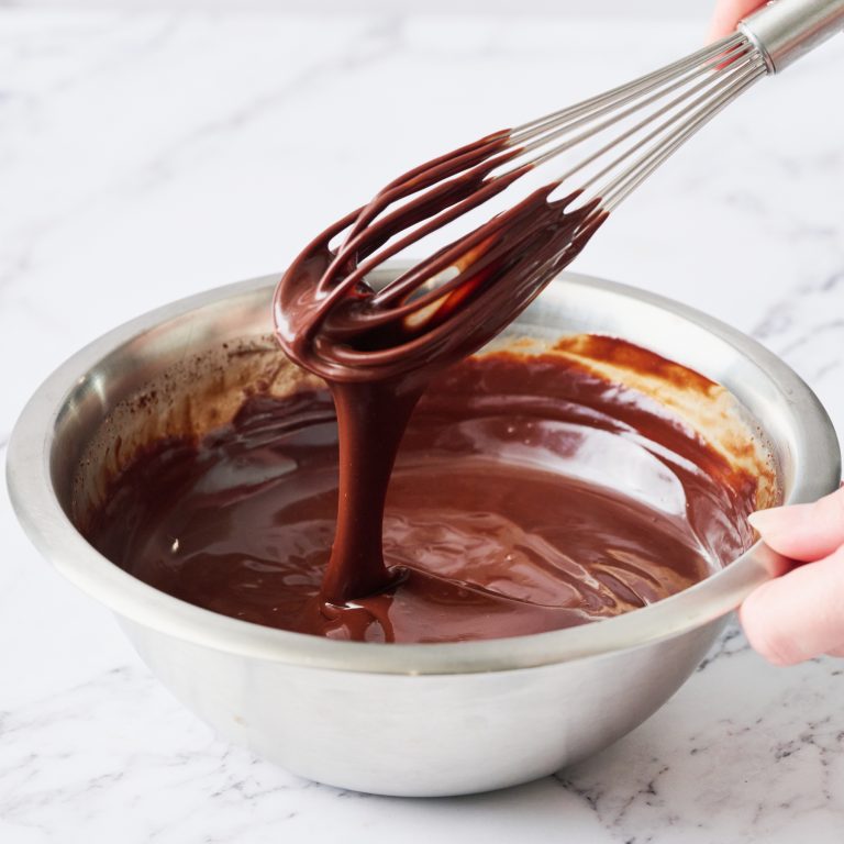 a bowl of chocolate ganache on a marble surface, with a hand holding a whisk being pulled out of the shiny ganache.