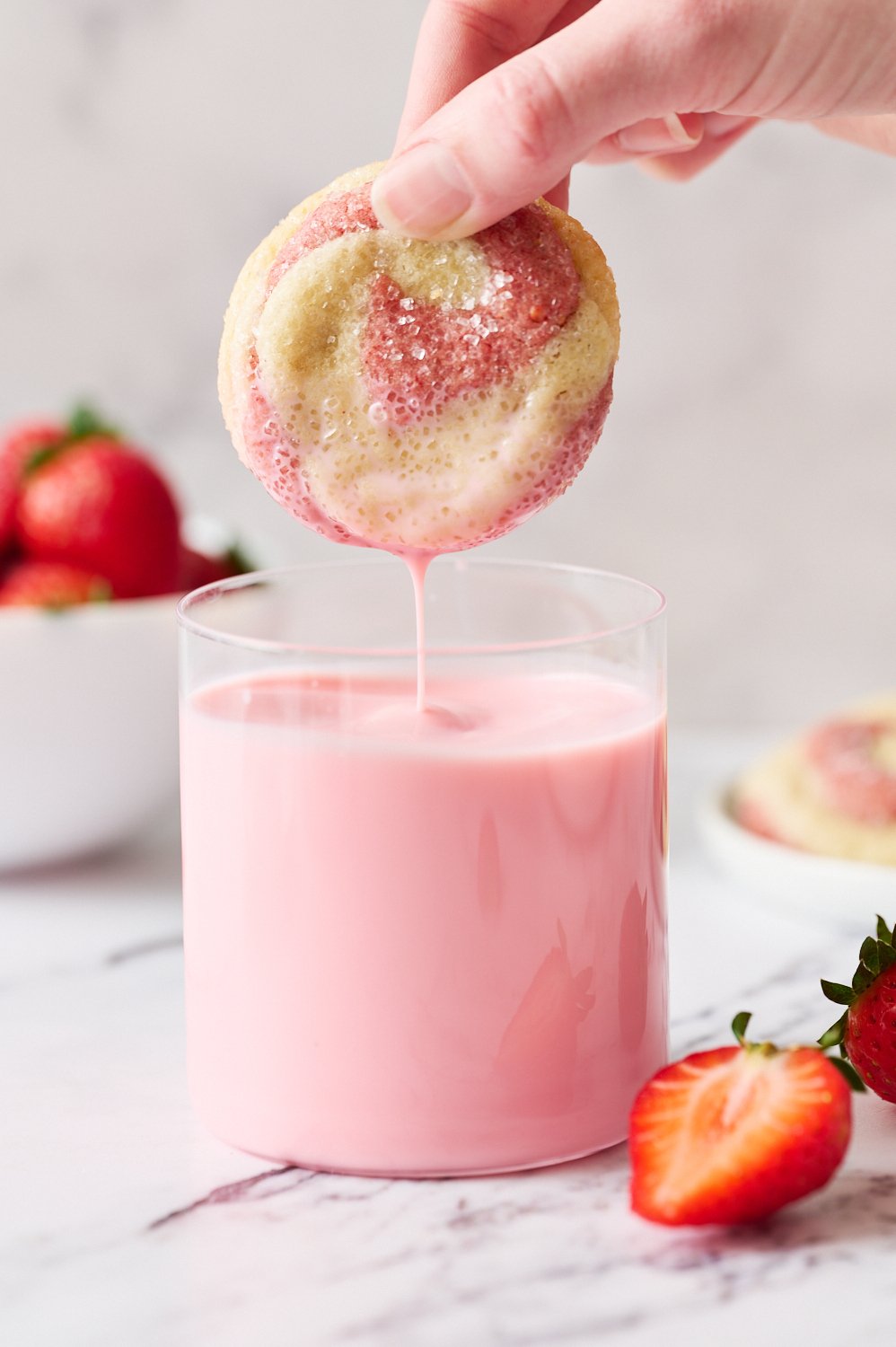a hand dunking a strawberry swirl sugar cookie in a glass of strawberry milk.