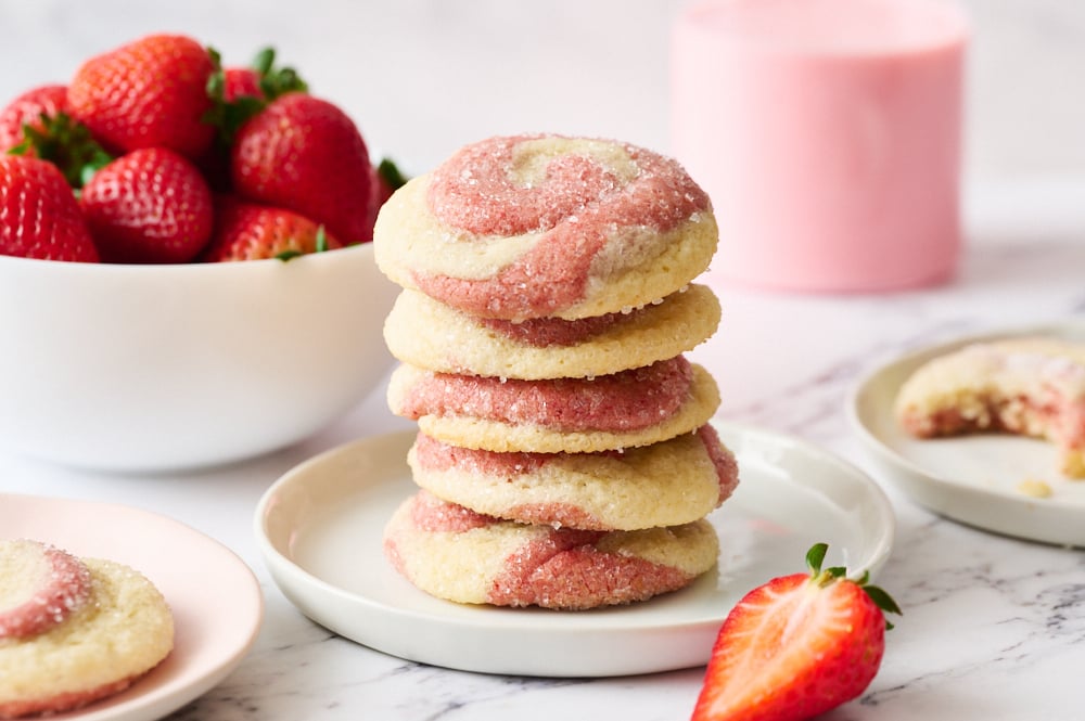 a stack of strawberry swirl sugar cookies on a plate, with berries and a glass of strawberry milk in the background.
