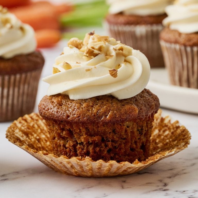 a carrot cake cupcake with its wrapper removed, iced with cream cheese frosting and sprinkled with some crushed walnuts on top.