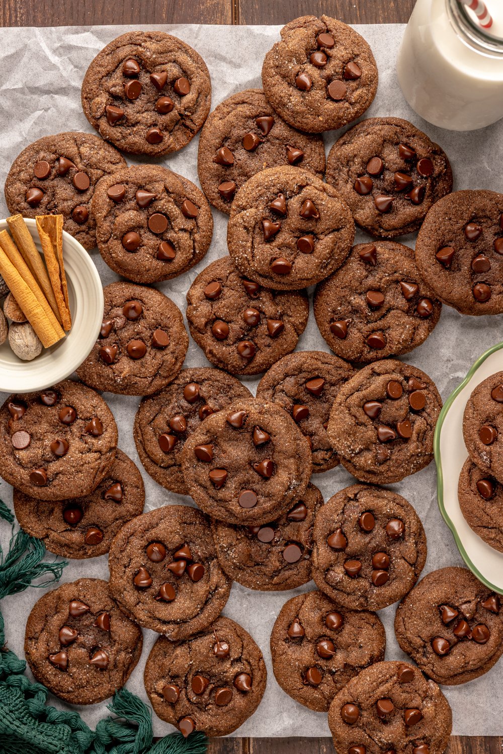 many cookies spread across a piece of parchment, with a bowl of spices and a glass of milk