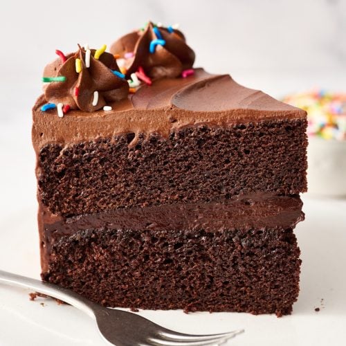 slice of tall chocolate cake on a plate with a fork and a small bowl of rainbow sprinkles in the background