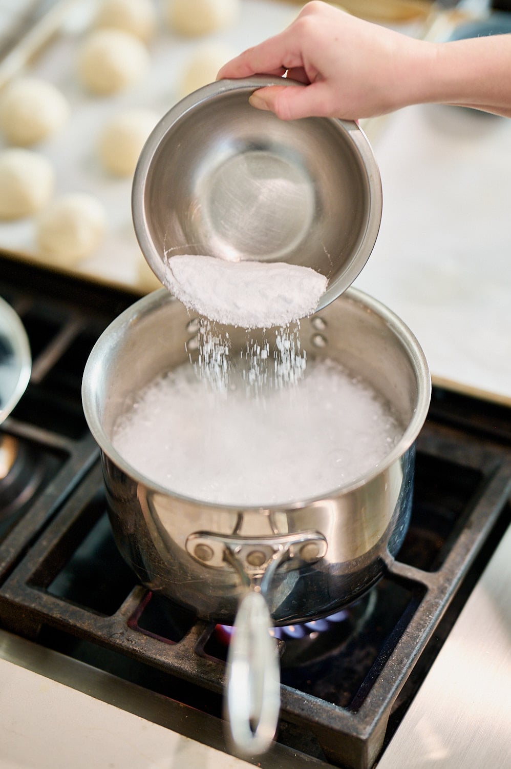 view of saucepan filled with boiling water on stovetop, a hand pouring baking soda from a bowl into saucepan