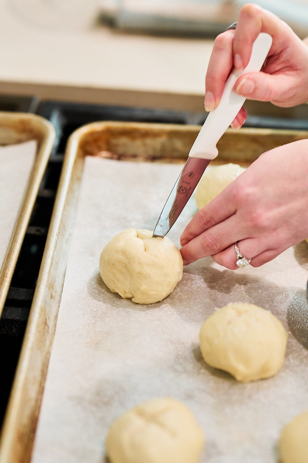 hand holding knife scoring a criss cross pattern into a bathed pretzel bun on a parchment lined pan