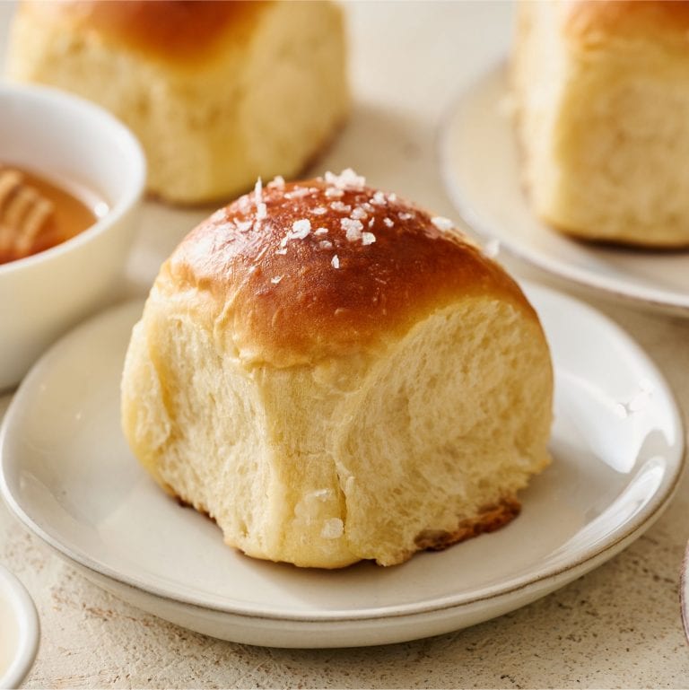 a small bread plate with a honey butter roll on top, a small bowl of honey with a honeycomb behind it, and more plated rolls