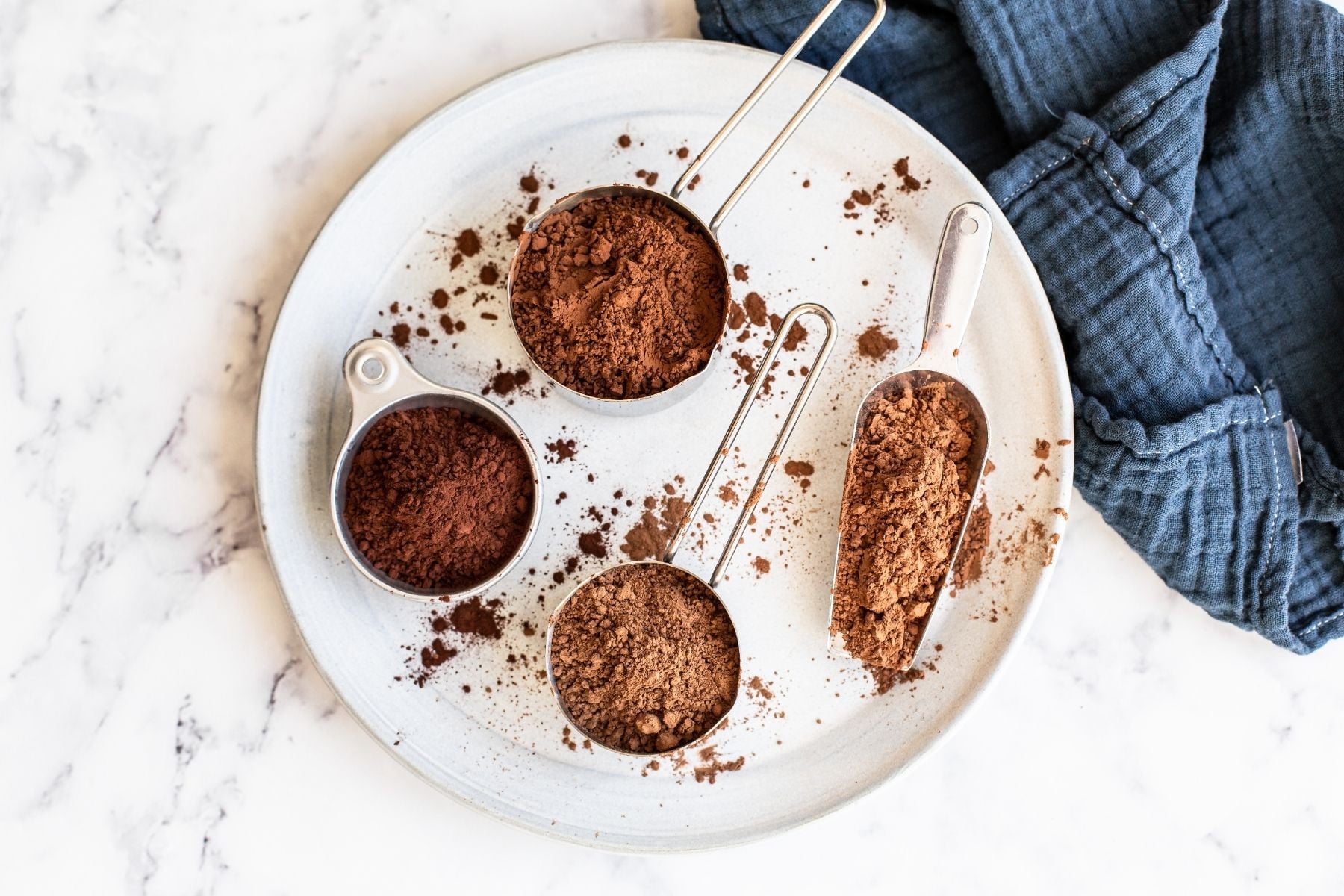 a large white plate with four measuring cups filled with different styles and brands of cocoa on a marble counter with a dark blue napkin