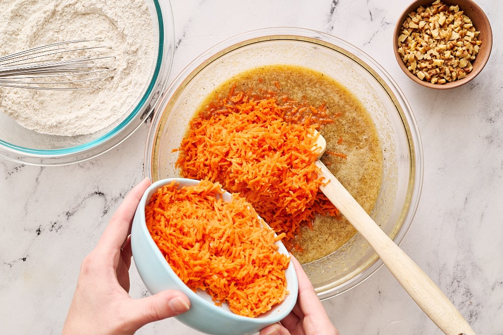 adding freshly grated carrots to a glass mixing bowl containing all the wet ingredients