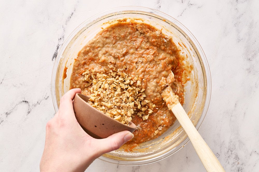 pouring nuts into carrot cake batter in glass mixing bowl