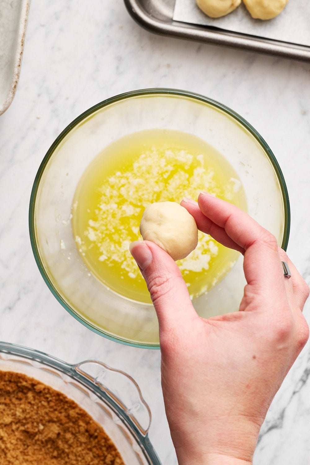 A hand holding a small round of dough, about to dip into a small glass bowl of melted butter