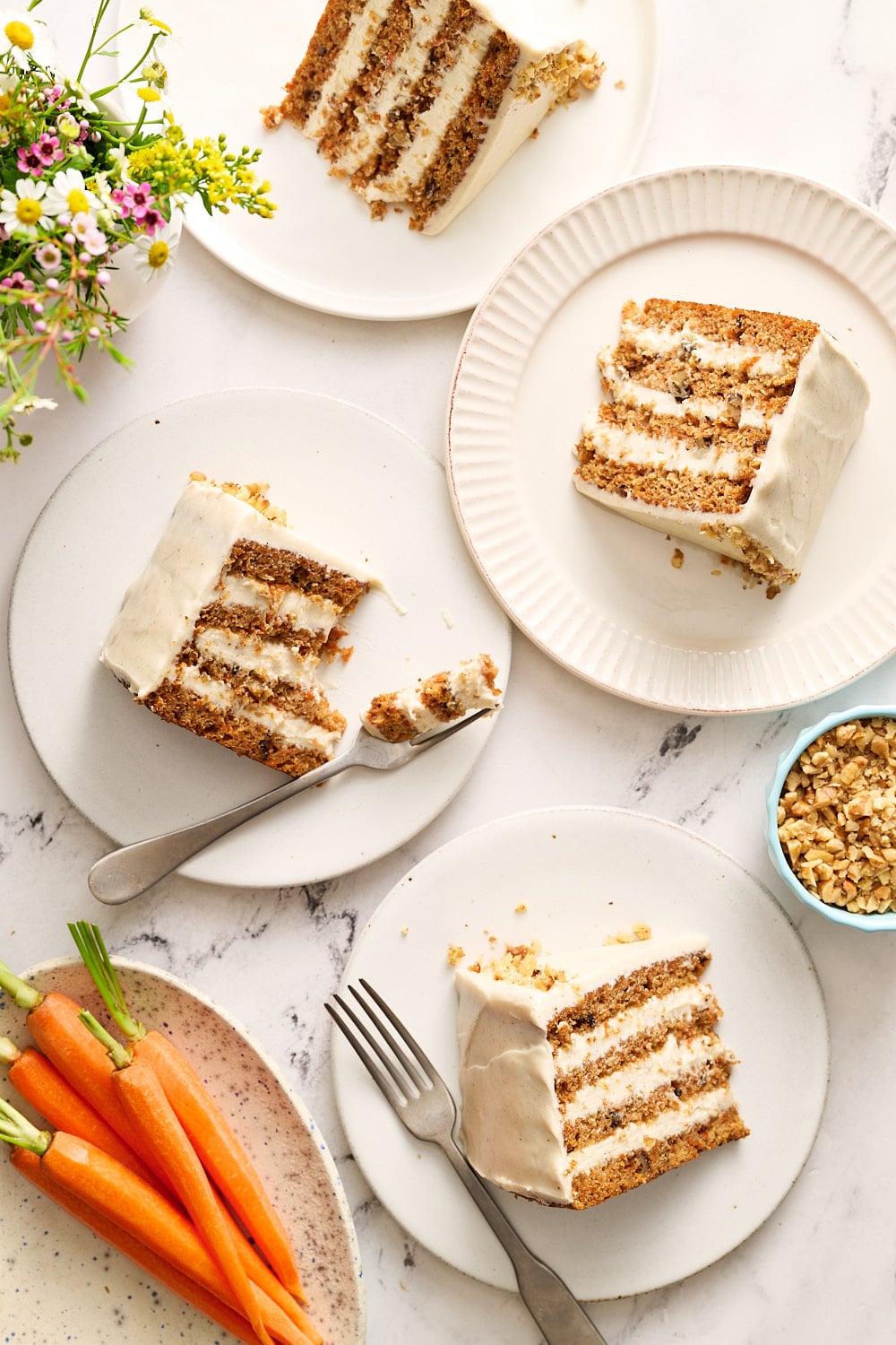several slices of carrot cake on plates on a marble surface with flowers and fresh carrots