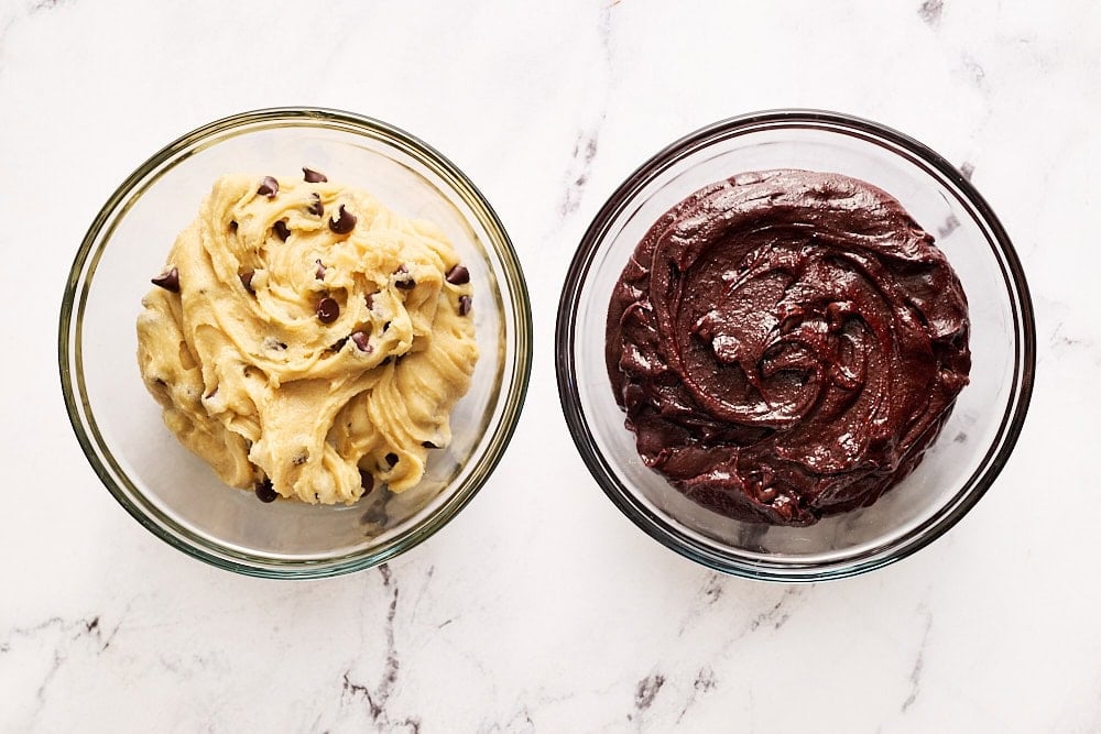 A glass bowl of cookie dough and a glass bowl of brownie batter on a marble counter