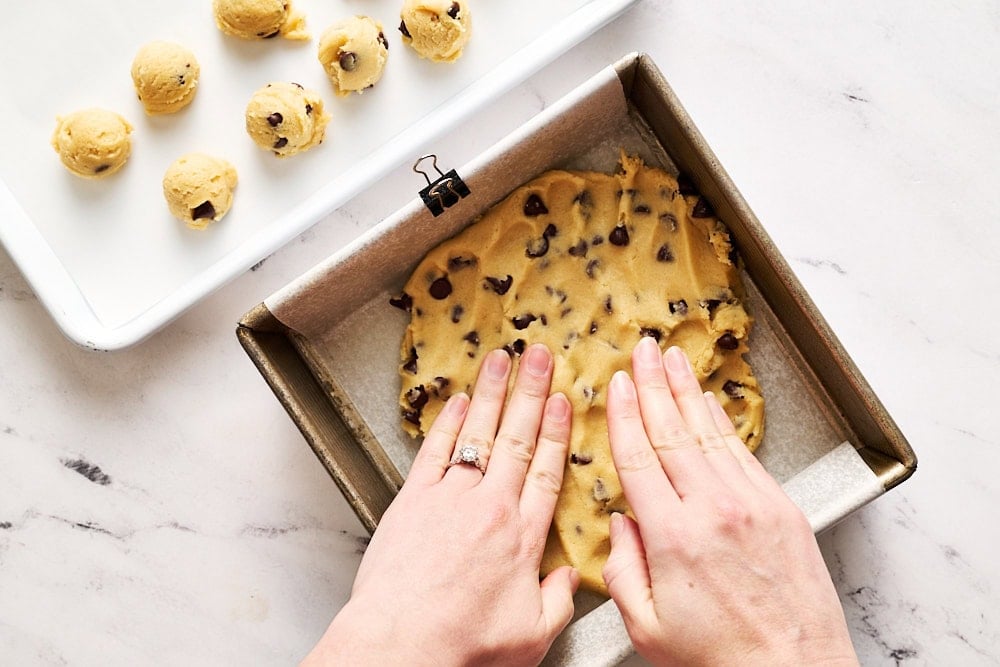 pressing cookie dough into a square metal pan, with more cookie dough on a tray shaped into small balls