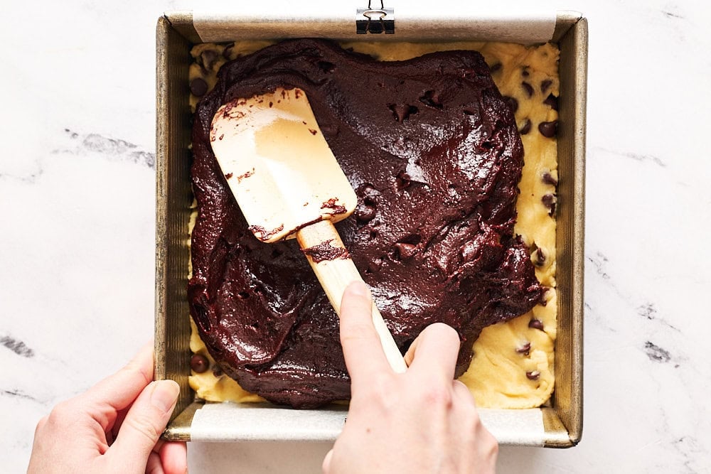 spreading shiny brownie batter on top of the cookie dough layer with a white rubber spatula on a marble counter