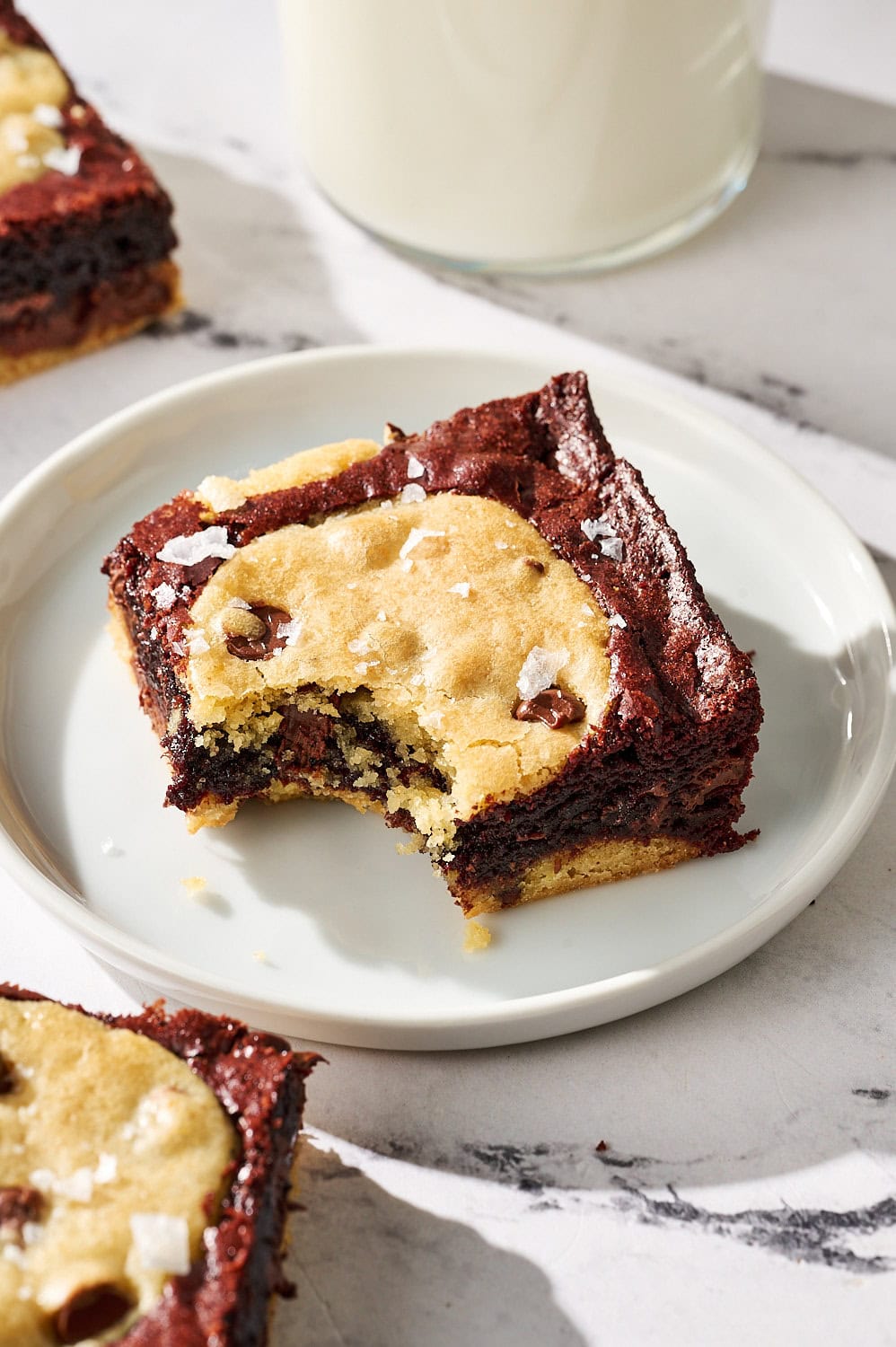a fudgy brookie with a bite taken out on a small white plate on a marble counter, with a glass of milk
