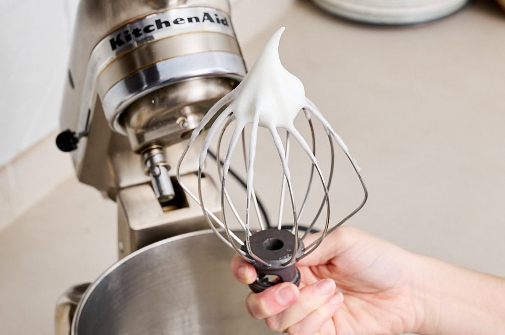 a hand holding the whisk attachment from a stand mixer showing egg whites whipped to soft peaks