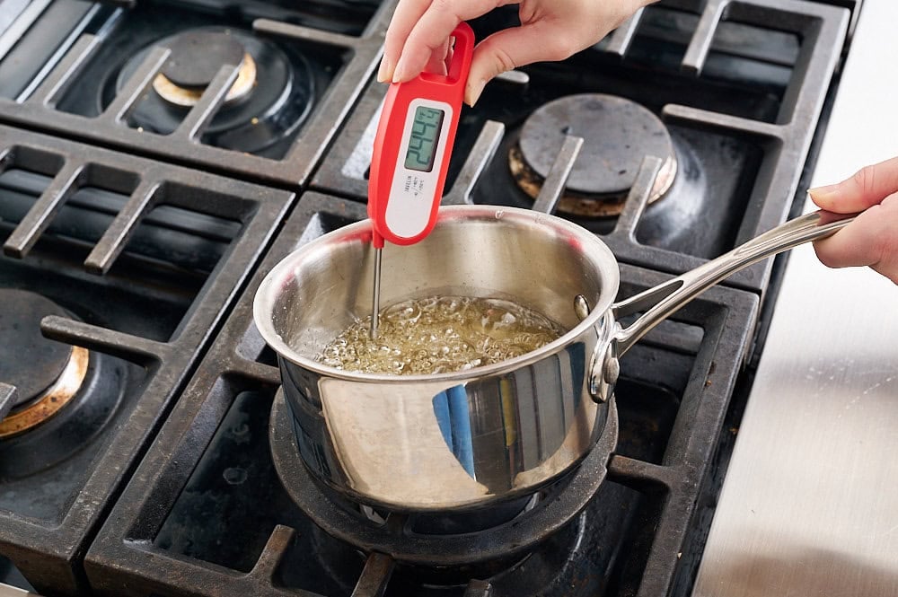 a small saucepan on a stovetop cooking the syrup mixture with another hand measuring the temperature with an instant read thermometer