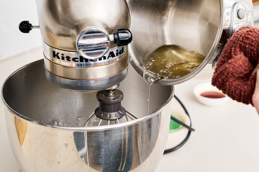 a saucepan slowing streaming in the syrup mixture into the stand mixer bowl with the whipped egg whites