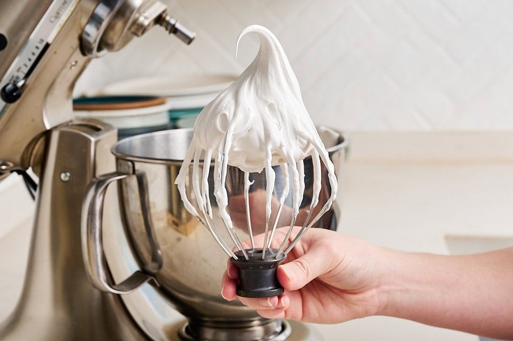 a hand holding the whisk attachment showing a tall shiny peak of the final marshmallow fluff