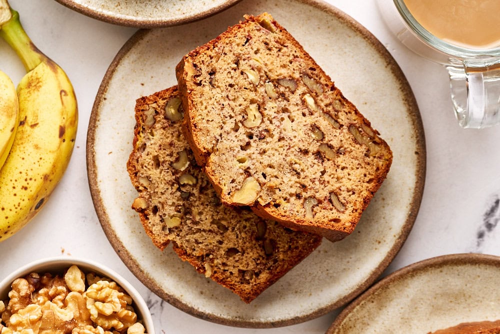 overhead view of two slices of banana bread with nuts on a plate, with a mug of coffee, a small bowl of extra nuts, and some extra bananas on a marble surface