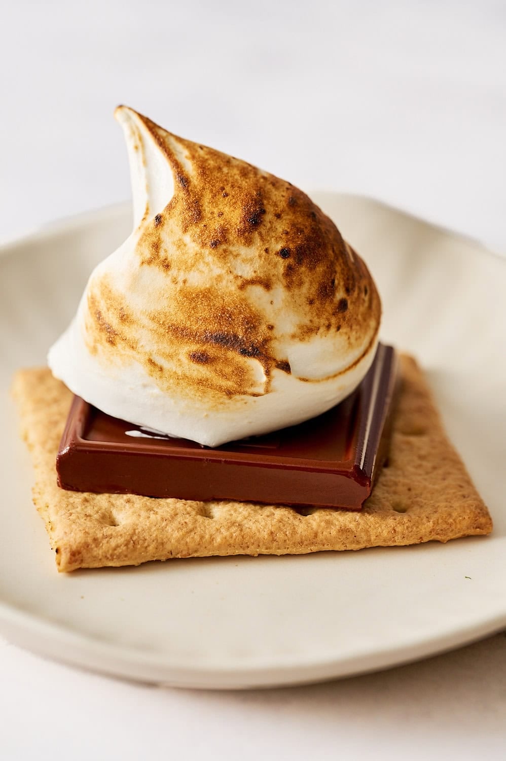 a graham cracker on a small plate with a piece of chocolate and a generous dollop of marshmallow creme that's been toasted on top