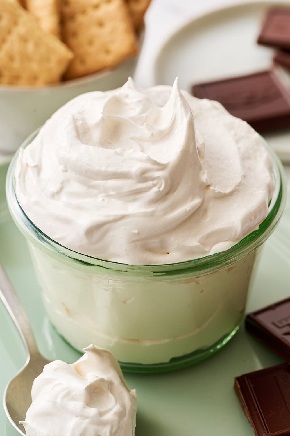 homemade marshmallow fluff in a glass container on a plate with a spoonful and chocolate squares and a bowl of graham crackers in the background