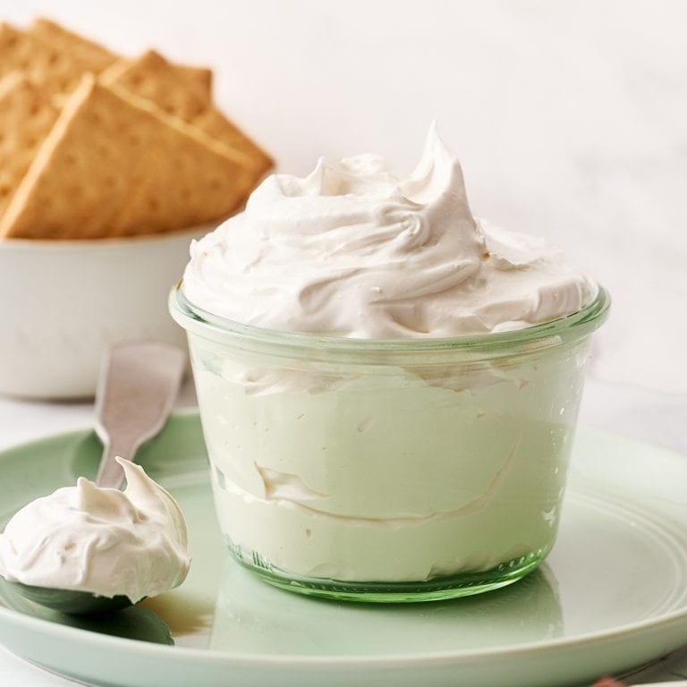 homemade marshmallow fluff in a glass container on a plate with a spoonful and a bowl of graham crackers in the background