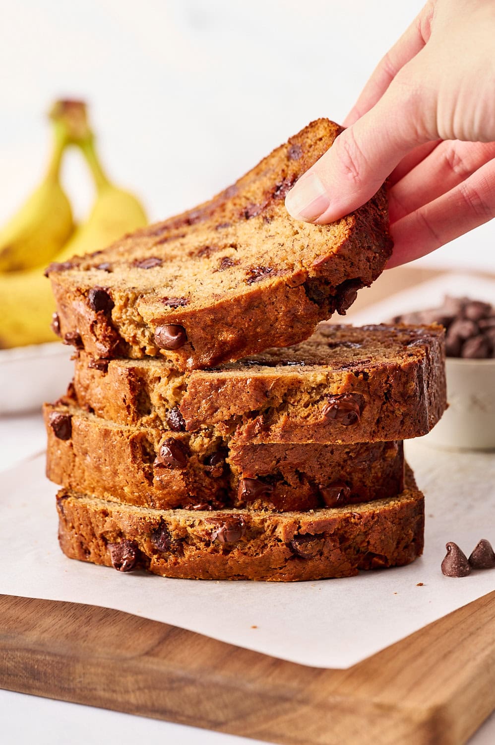 stack of banana bread slices with chocolate chips on a parchment lined wooden board, with a hand removing the top slice